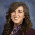 headshot of a smiling woman with curly light brown hair dressed in a maroon blazer and blue and pink floral shirt